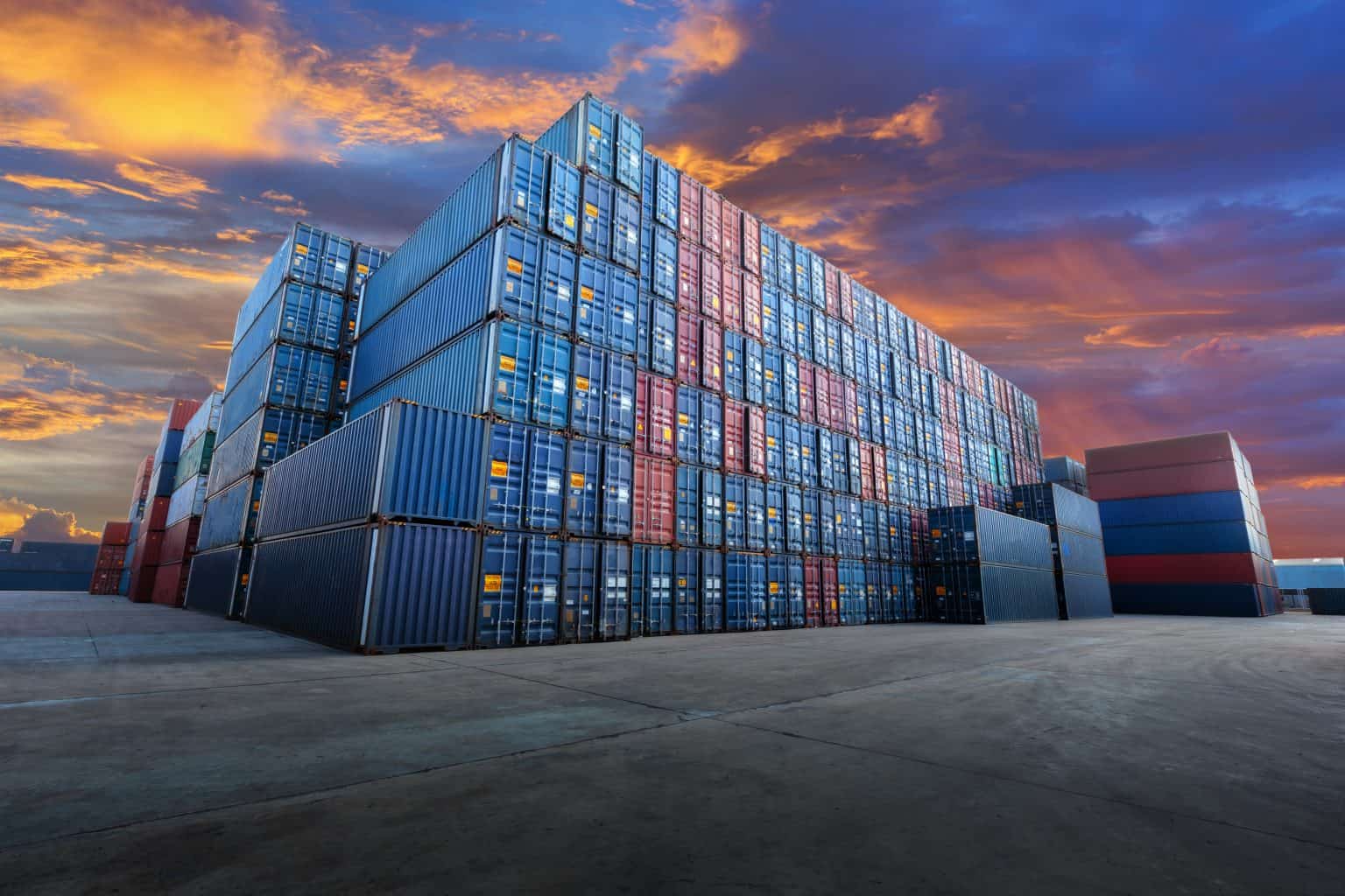 A large stack of high-quality, colorful shipping containers is arranged systematically at a busy port yard under a vivid, dramatic sunset sky. The vibrant hues of the containers contrast with the serene, expansive concrete ground, creating a visually striking industrial scene.